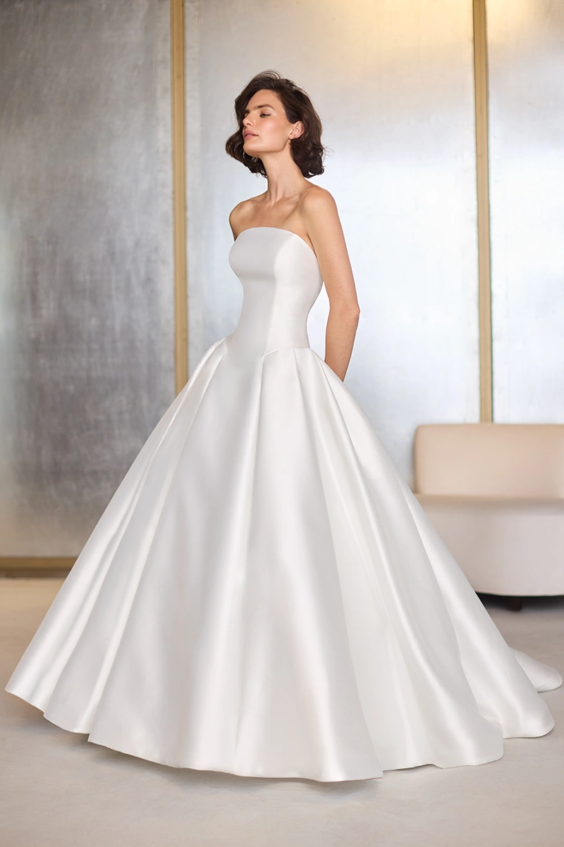 Arched neckline Mikado princess cut gown with a strapless drop waisted bodice, a box pleated ball gown skirt, and a removable long sleeve sheer Lace bolero.