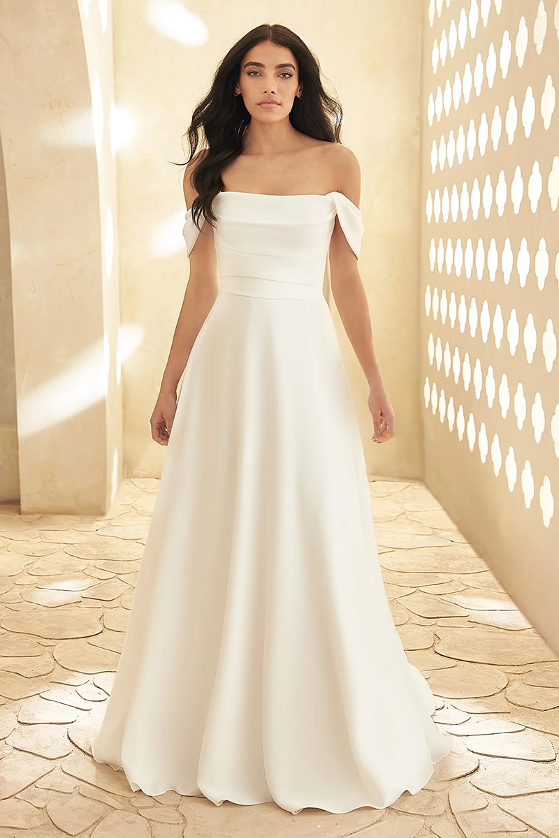 Off-shoulder crepe wedding gown with a folded neckline, and flowing A-line skirt shown in textured architectural setting.
