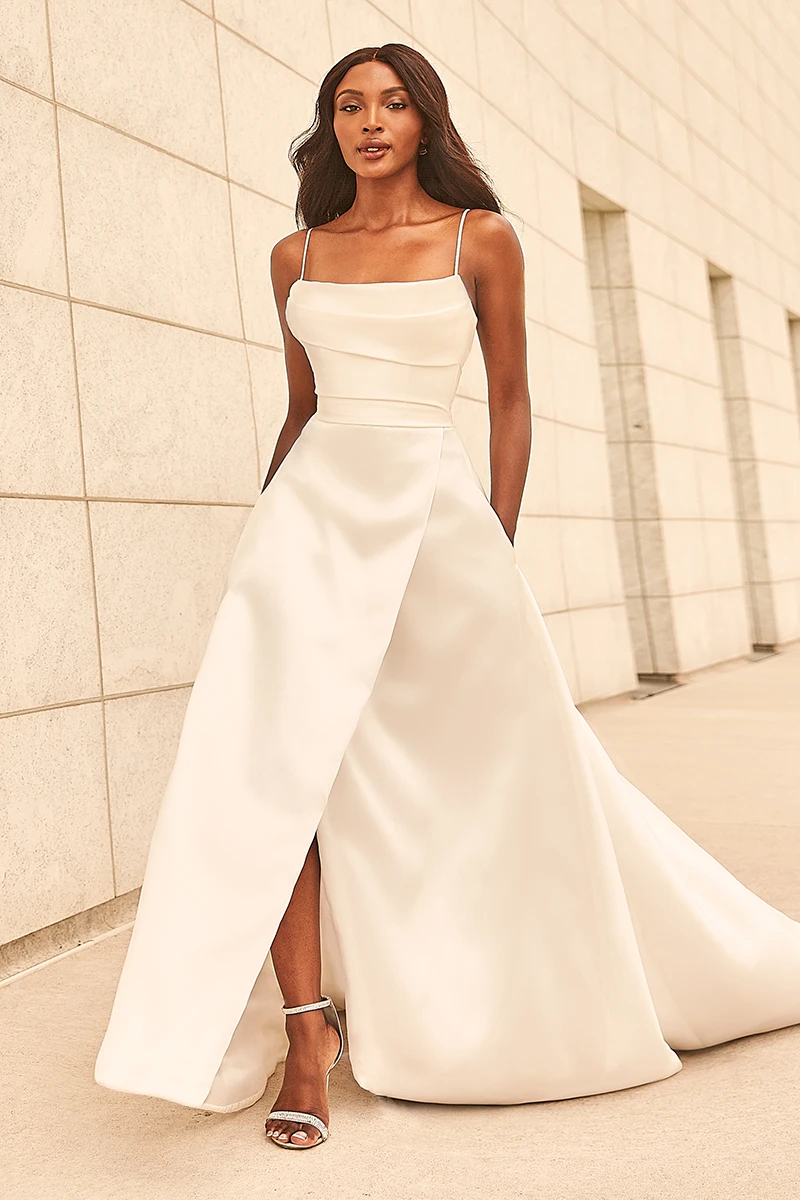 A-line wedding gown with spaghetti straps, a scoop neckline, and front skirt slit shown in a white architectural setting.