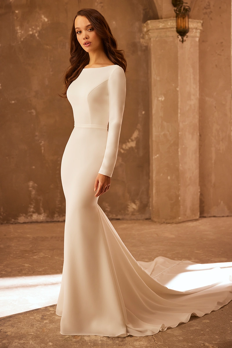 Elegant long-sleeve crepe wedding dress with a bateau neckline and chapel train, shown in a rustic indoor setting.