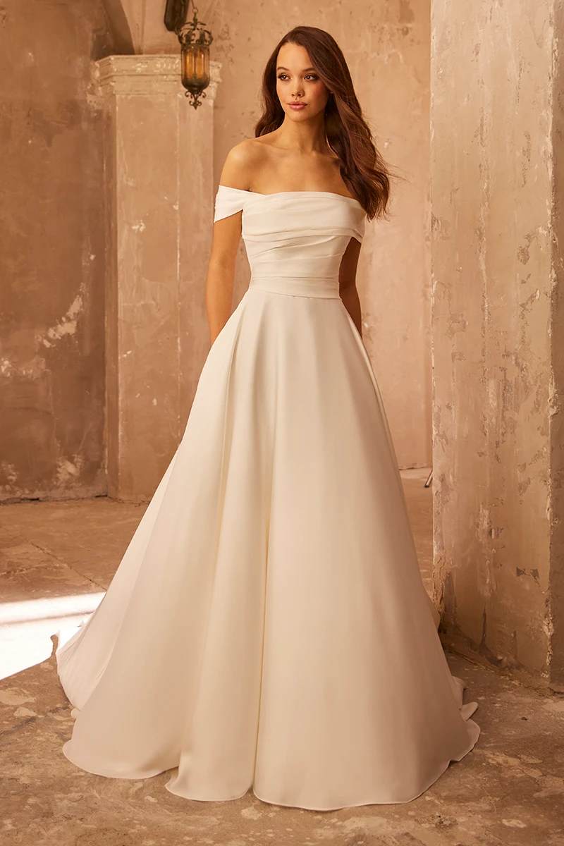 Off-shoulder A-line wedding dress with ruched satin bodice and flowing skirt, shown in a textured stone setting.