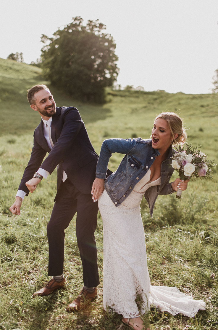 Bride laughing and dancing in a fitted lace wedding gown with a denim jacket, holds a bouquet next to groom, in sunny meadow.