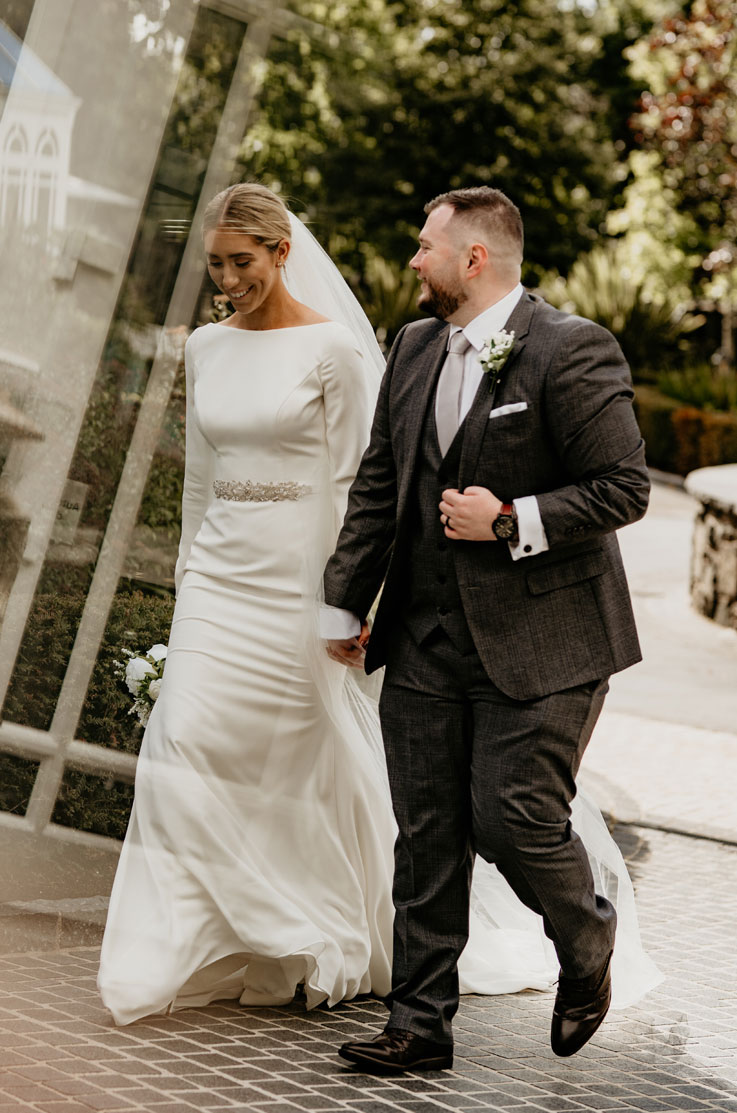 Bride in a crepe bateau neck gown with long sleeves and a beaded belt, walks hand in hand with groom in a black suit outdoors.