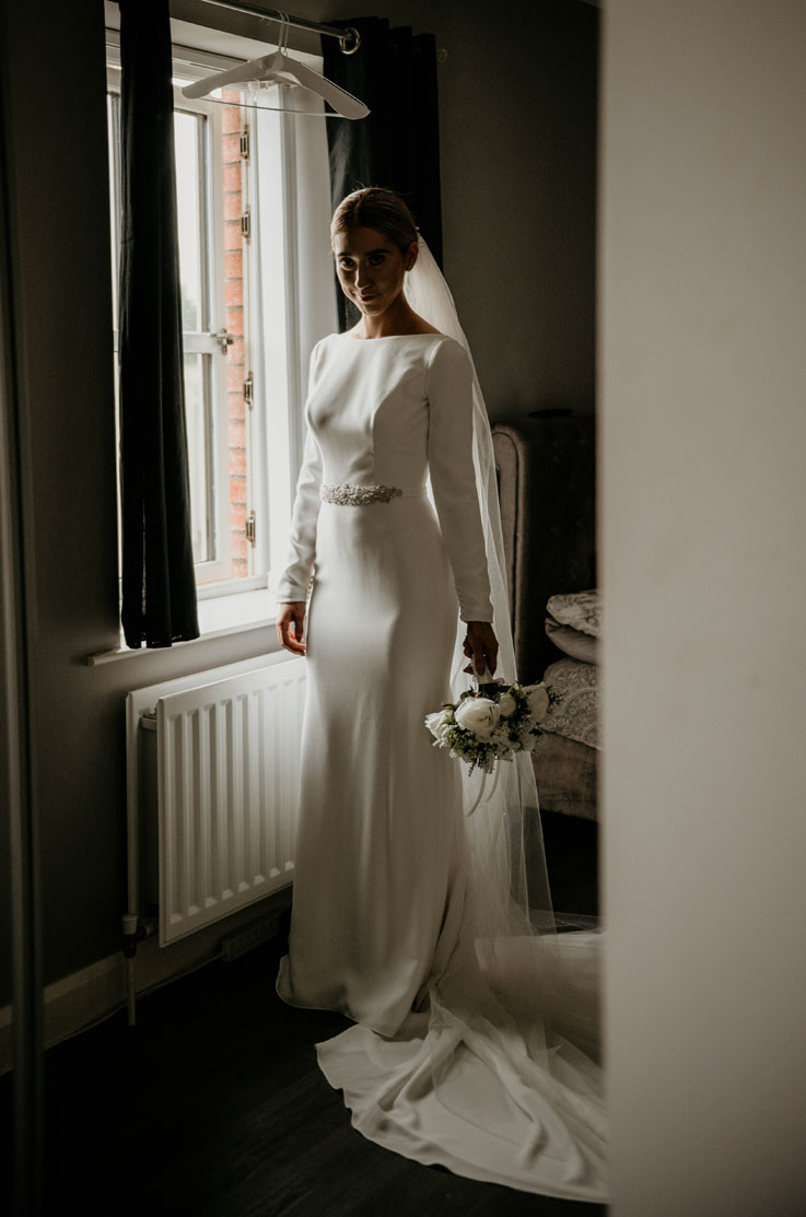 Bride stands near a bright open window in a high-neck crepe gown with long sleeves, belt and a veil, holding a bouquet.