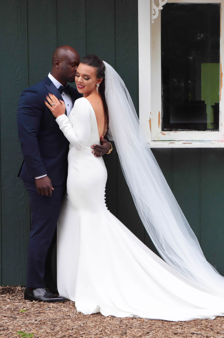 Groom embraces his bride in a backless long sleeve gown with a veil and buttoned train, in front of venue.
