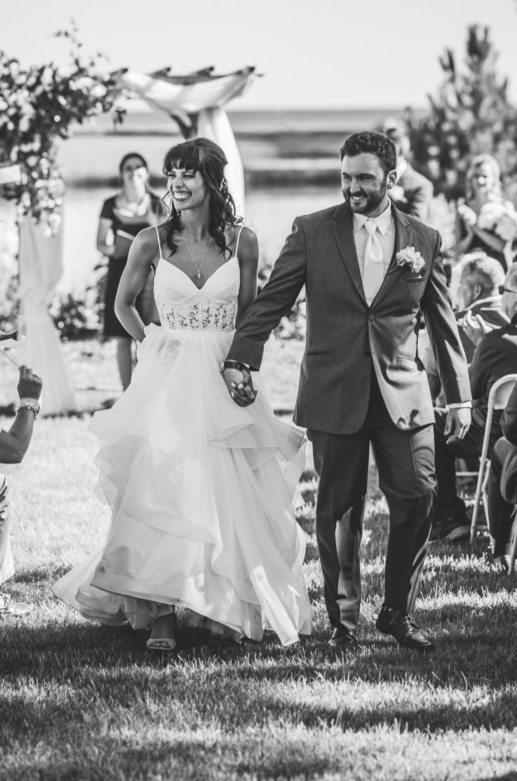 Bride in a lace and tulle wedding gown with a layered skirt, smiling and walks with groom in the aisle, in black and white.