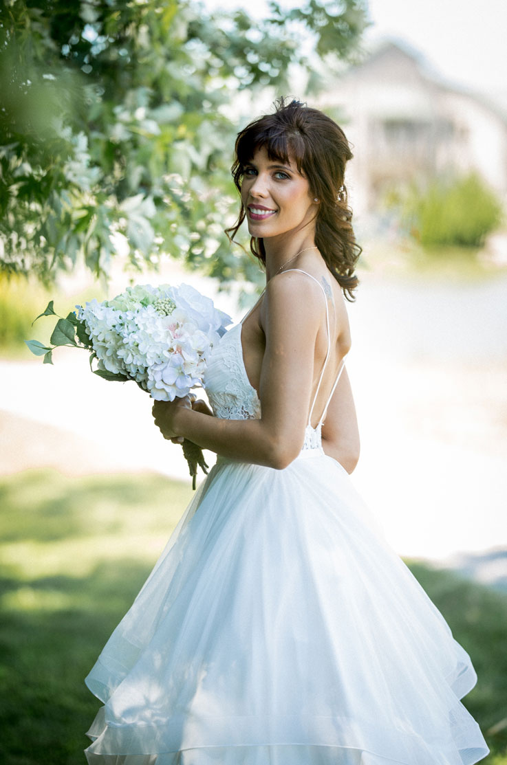 Close up of smiling bride in an backless lace bodice gown with a layered tulle skirt, holds a bouquet by a tree outside venue.