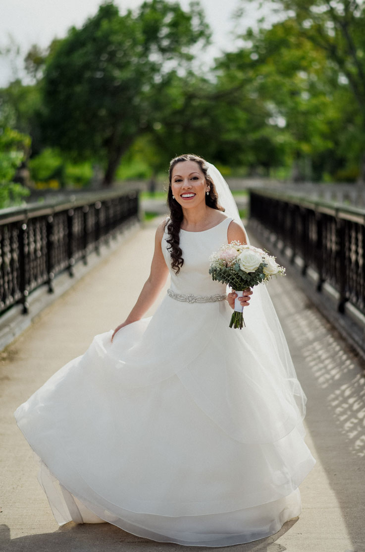  Smiling bride posing on a bridge surrounded by trees, in a satin and organza gown with a beaded belt, bateau neck and veil.