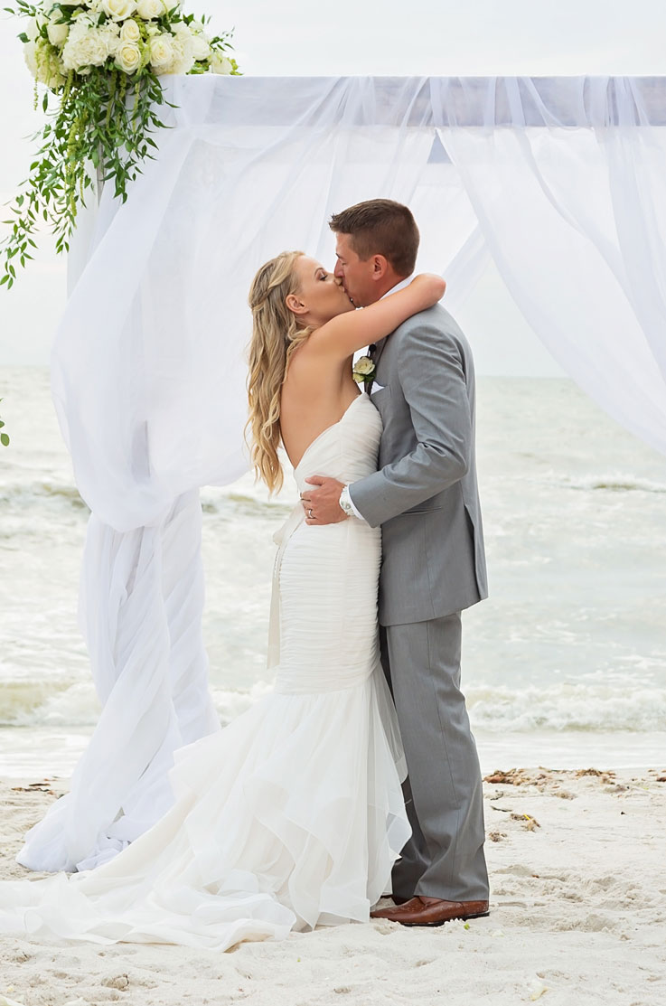 Bride in a backless tulle gown kisses her groom in a gray suit at altar, with flowing curtains at a shoreline with flowers.