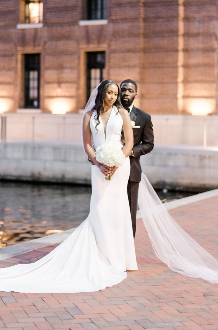 Groom embraces his bride in a sleek crepe gown with a halter plunging neckline and a veil, holding a bouquet by a waterfront.