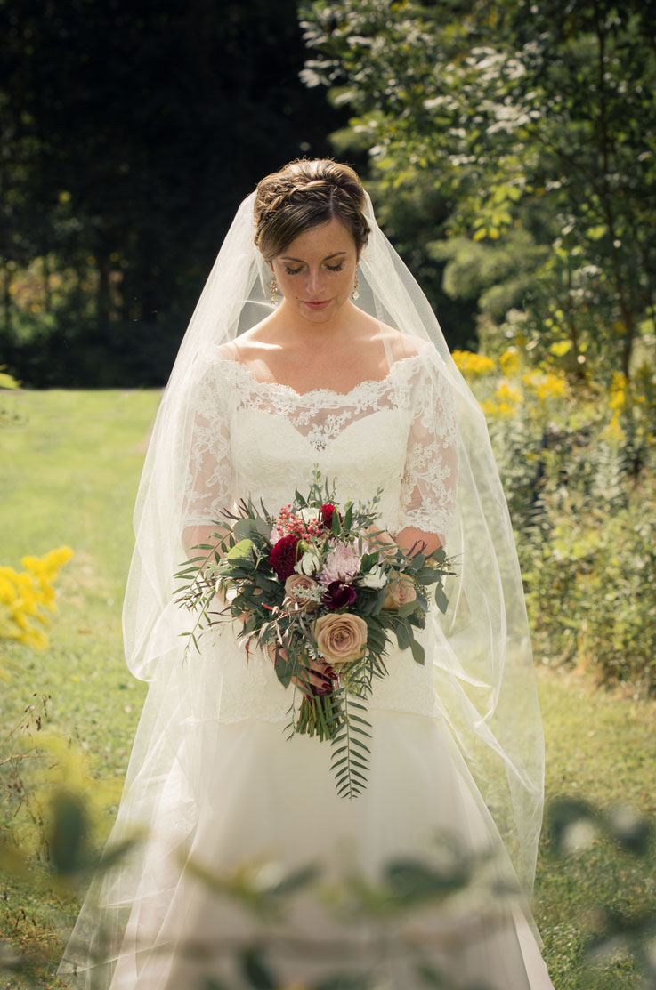 Close-up of bride in a lace and organdy gown, with an A-line skirt and veil, holds a bouquet in an open field.
