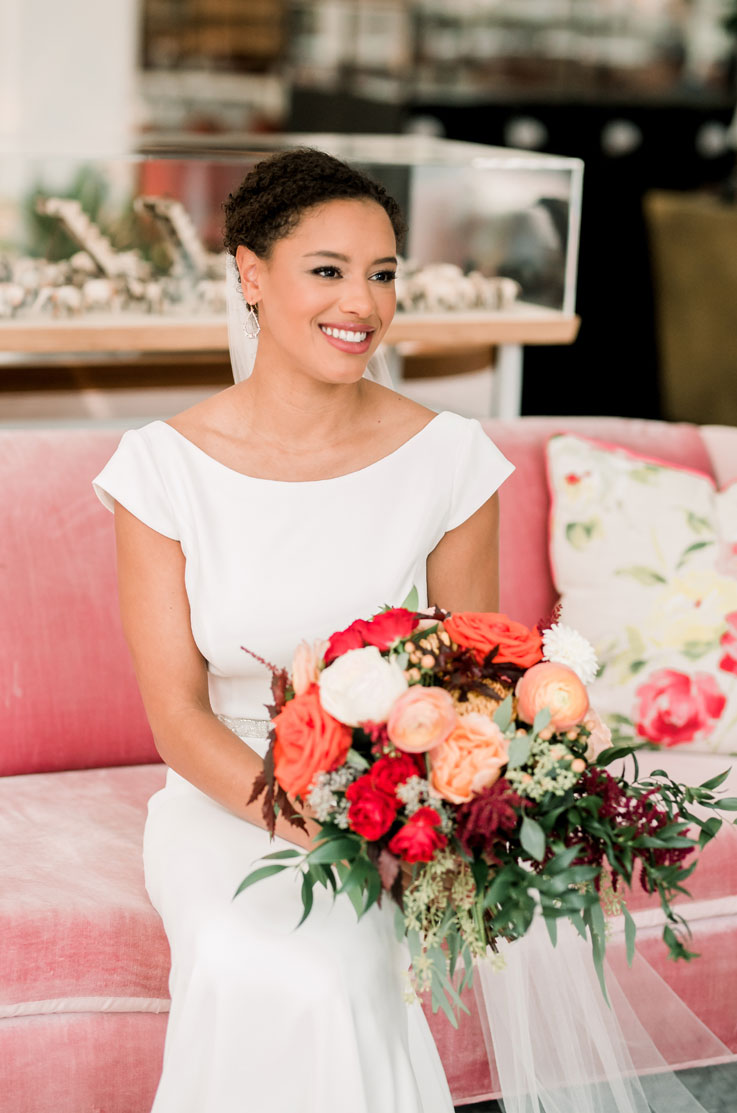  Smiling bride in a cap sleeve crepe gown with a veil and boat neckline, sits on a velvet couch indoors holding a bouquet.