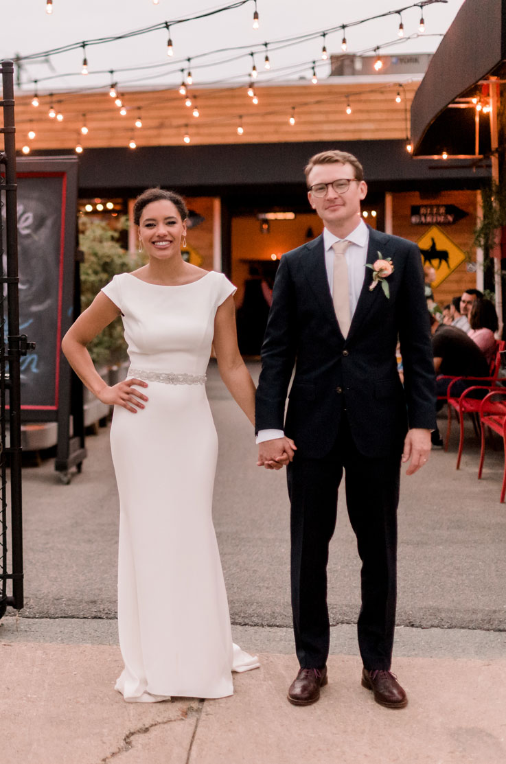 Bride poses outside venue holding hands with groom and wearing a crepe fit and flare gown with a belt and cap sleeves, under outdoor string lights.