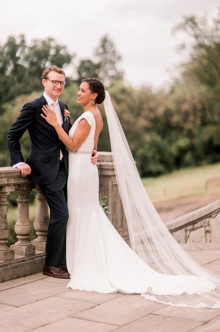 Bride in a backless crepe gown with a flowing veil, belt and cap sleeves, smiles at groom leaning on a stone railing on a terrace.