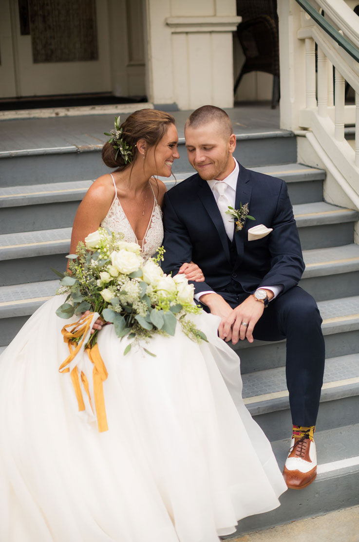 Bride in a V-neck lace gown with tulle, sits on steps in front of the venue, beside groom and smiling holding a rose bouquet.
