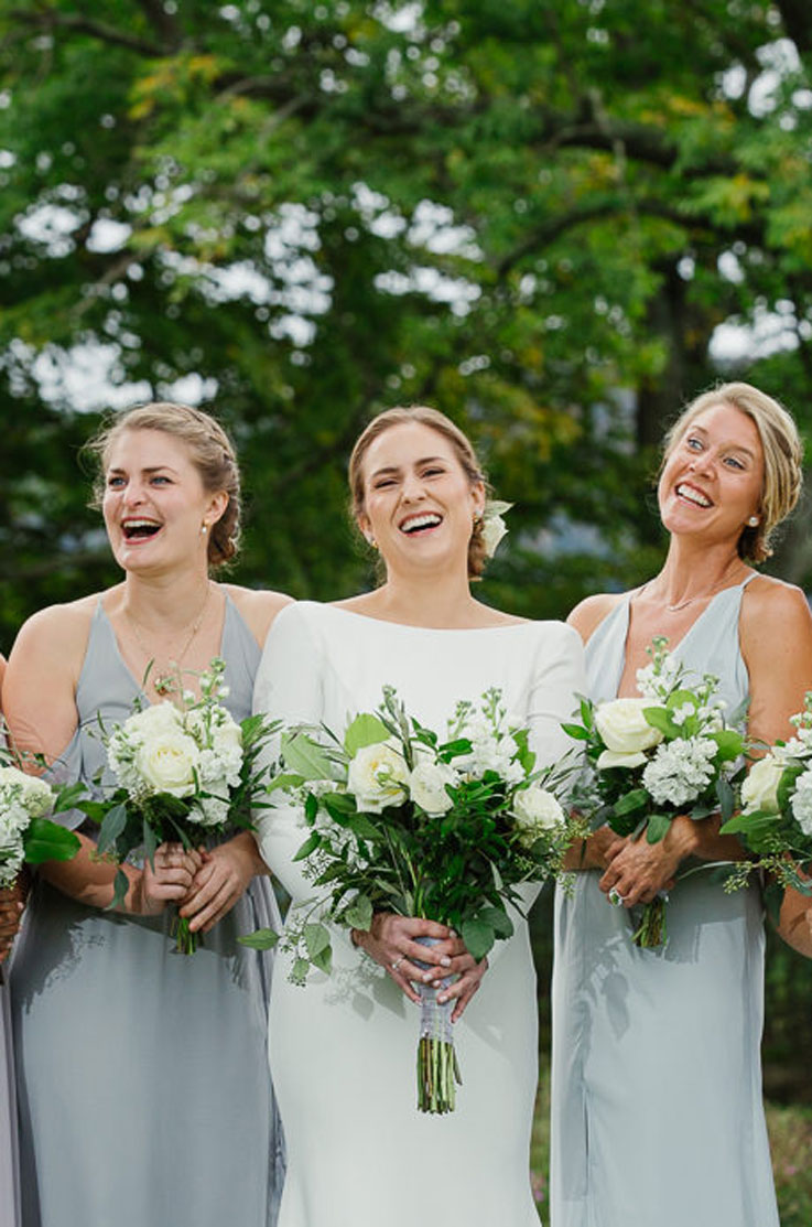 Bride in a long sleeve crepe gown with a bateau neckline, laughing with her bridesmaids in blue dresses and holding bouquets.