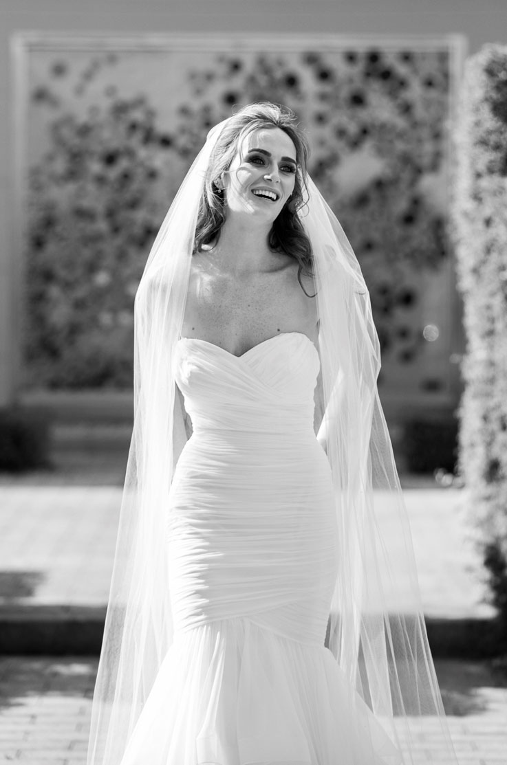 Smiling bride in a strapless wedding gown in tulle with a sweetheart neckline and veil, stands outdoors in black and white.