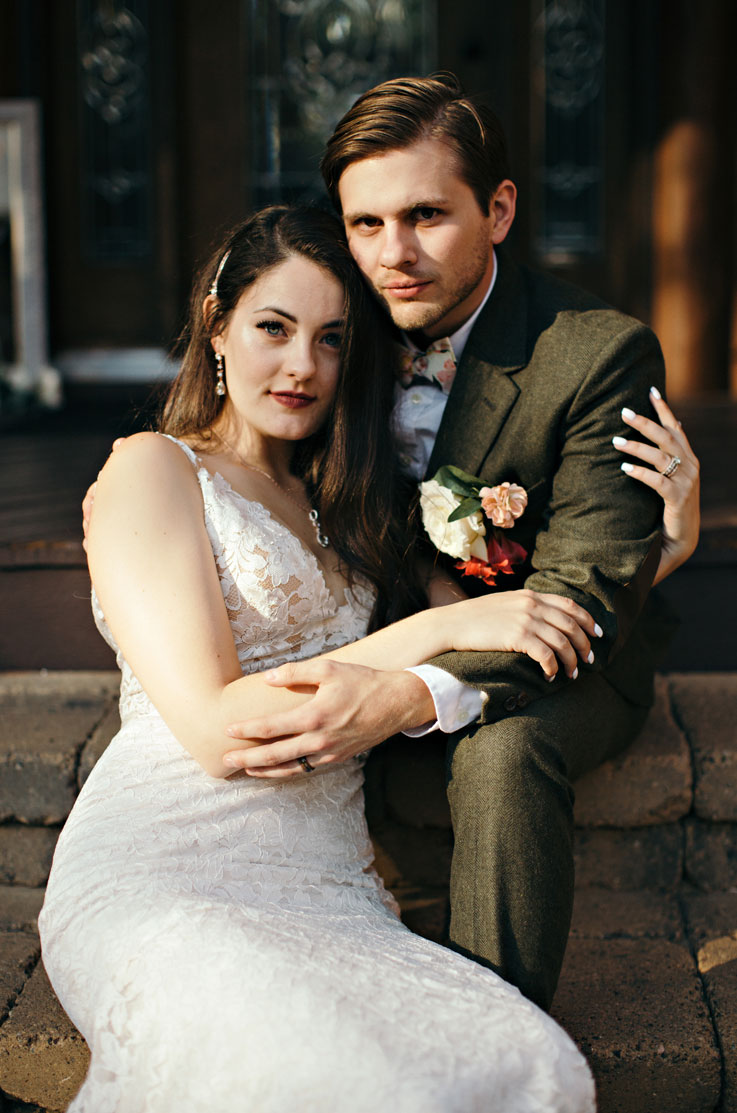 Groom poses with bride in a sleeveless lace wedding dress with floral designs and a V-neckline, sitting on stone steps.