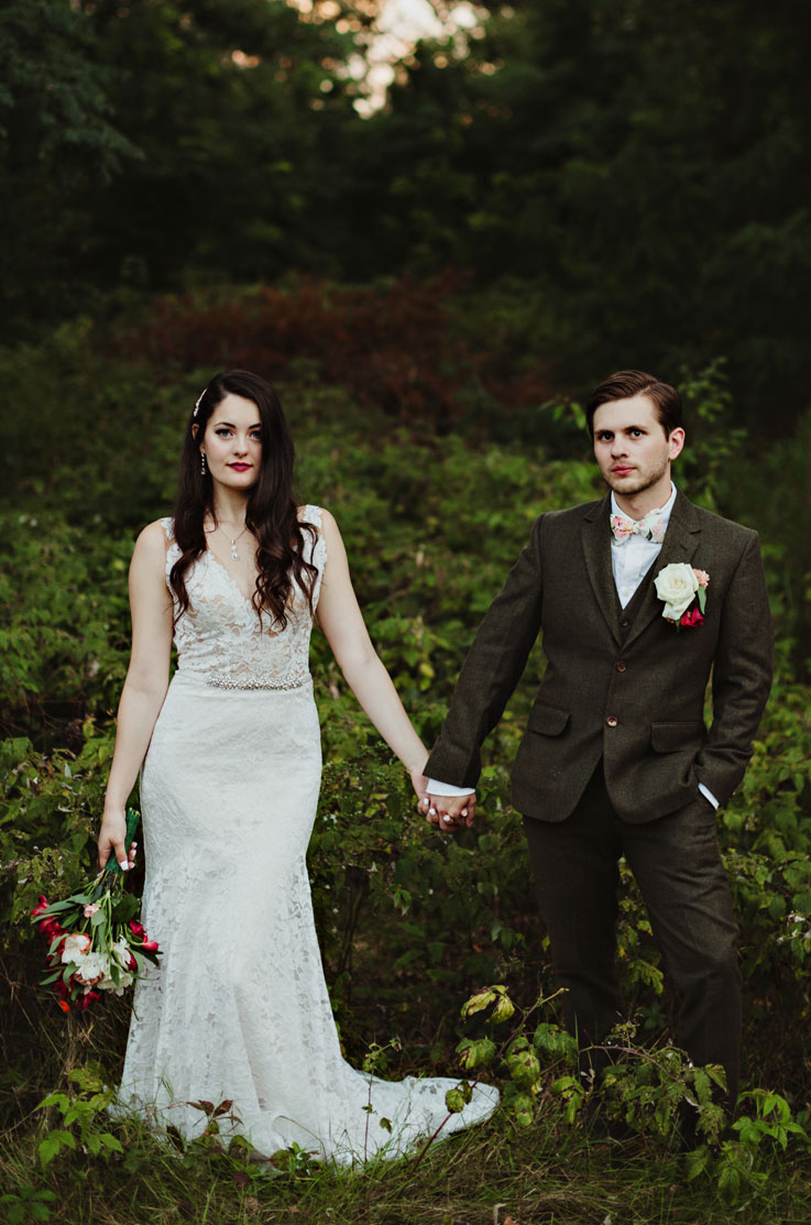 Bride in a lace fit and flare gown, holding hands with groom and a bouquet in the other hand, standing in front of bushes.Bride in a lace fit and flare gown, holding hands with groom and a bouquet in the other hand, standing in front of bushes.