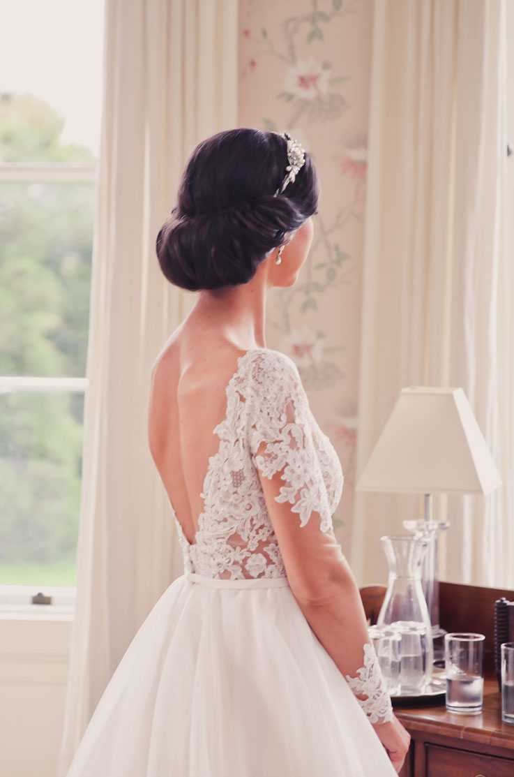 Back of bride in a lace long sleeved gown with a low open back and an updo, in an elegant interior with windows and a floral wall.