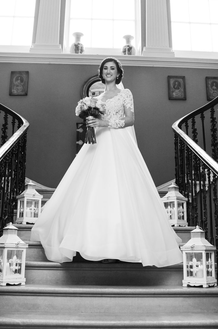 Bride in a lace and tulle A-line wedding dress with sleeves and a veil, standing on a staircase, shown in black and white.