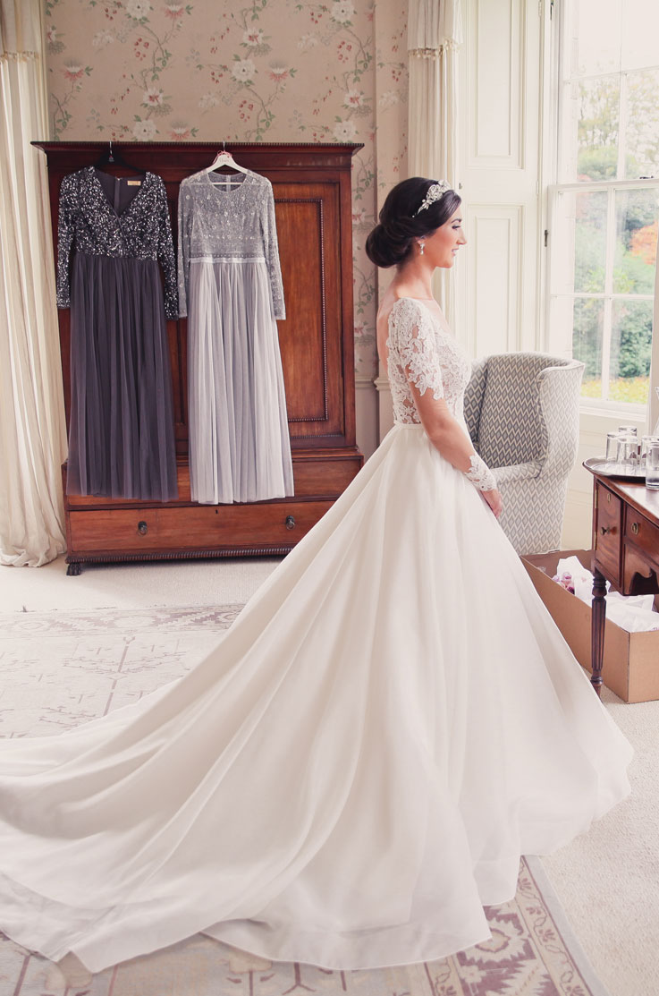 Bride in an illusion lace sleeve gown with tulle and a full skirt, wearing a headpiece and facing a window in a luxurious mansion.