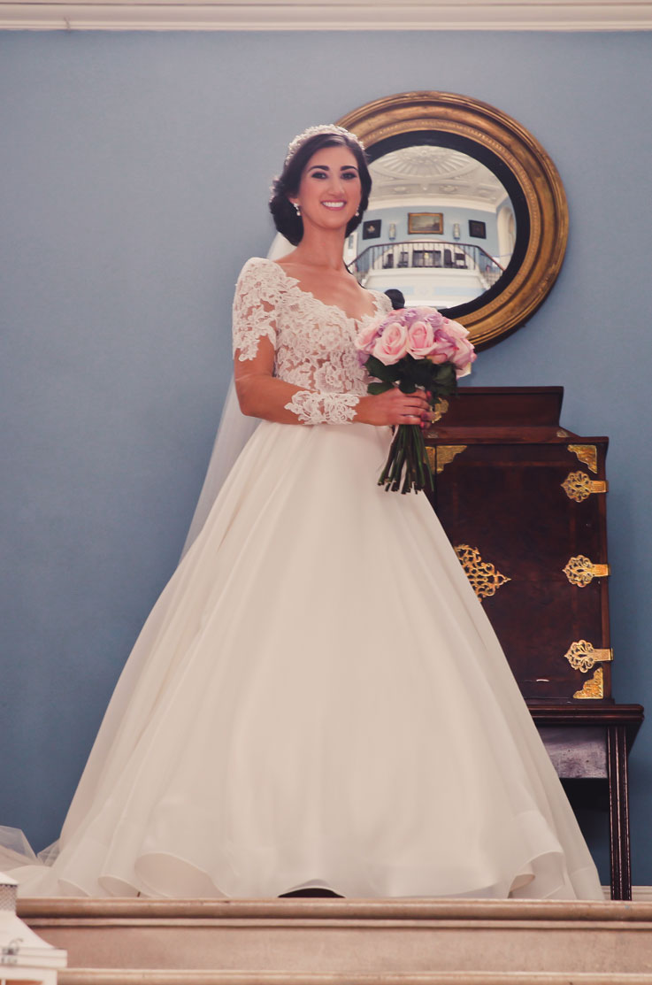 Smiling bride in a lace sleeve gown with an A-line organdy skirt and a flowing veil, holding a pink bouquet by a mirror.