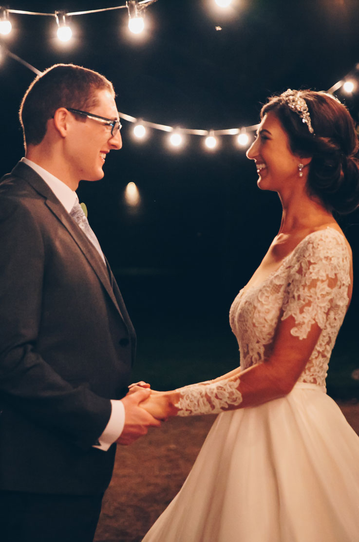 Groom faces and smiles at his bride in a lace bodice wedding gown with sleeves and a full skirt under string lights at night.