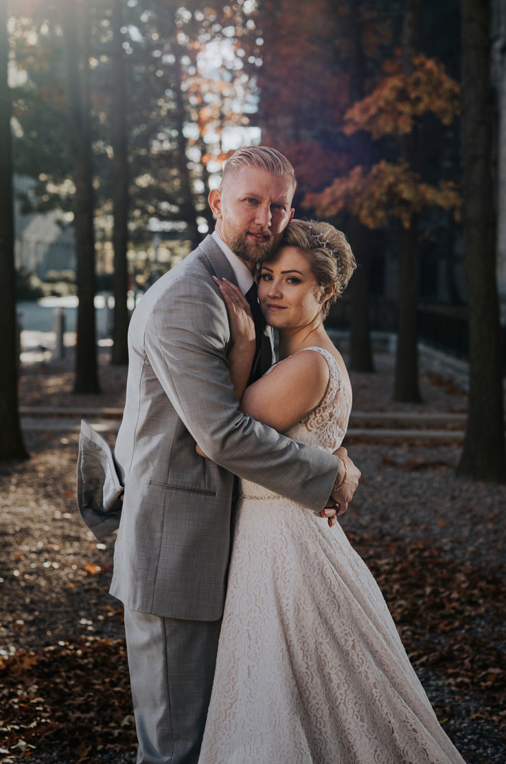 Groom holds bride in an embrace, wearing a sleevless lace gown with beaded belt detail, surrounded by autumn foliage.