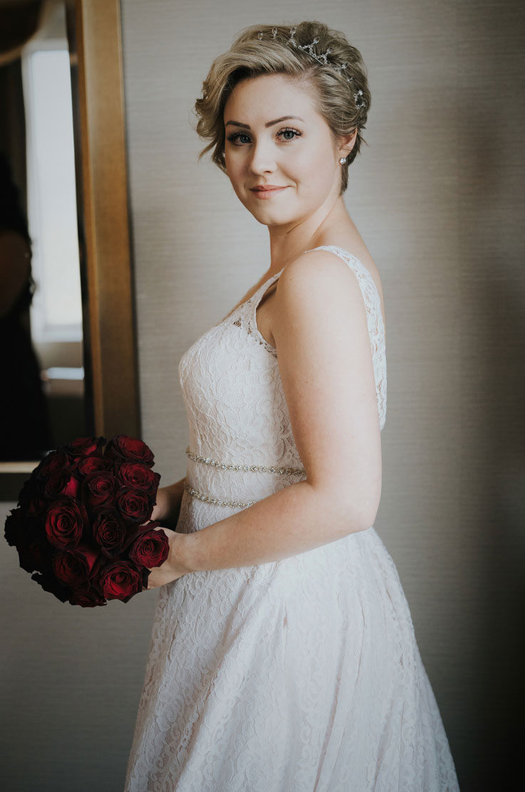  Close-up side view of a bride in a sleeveless lace gown with a double beaded belt, and is holding a dark rose bouquet in dark lighting.