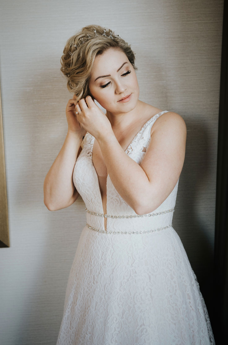 Close-up of bride getting ready indoors in a lace gown, with a plunging neckline and thin double beaded crystal belt.