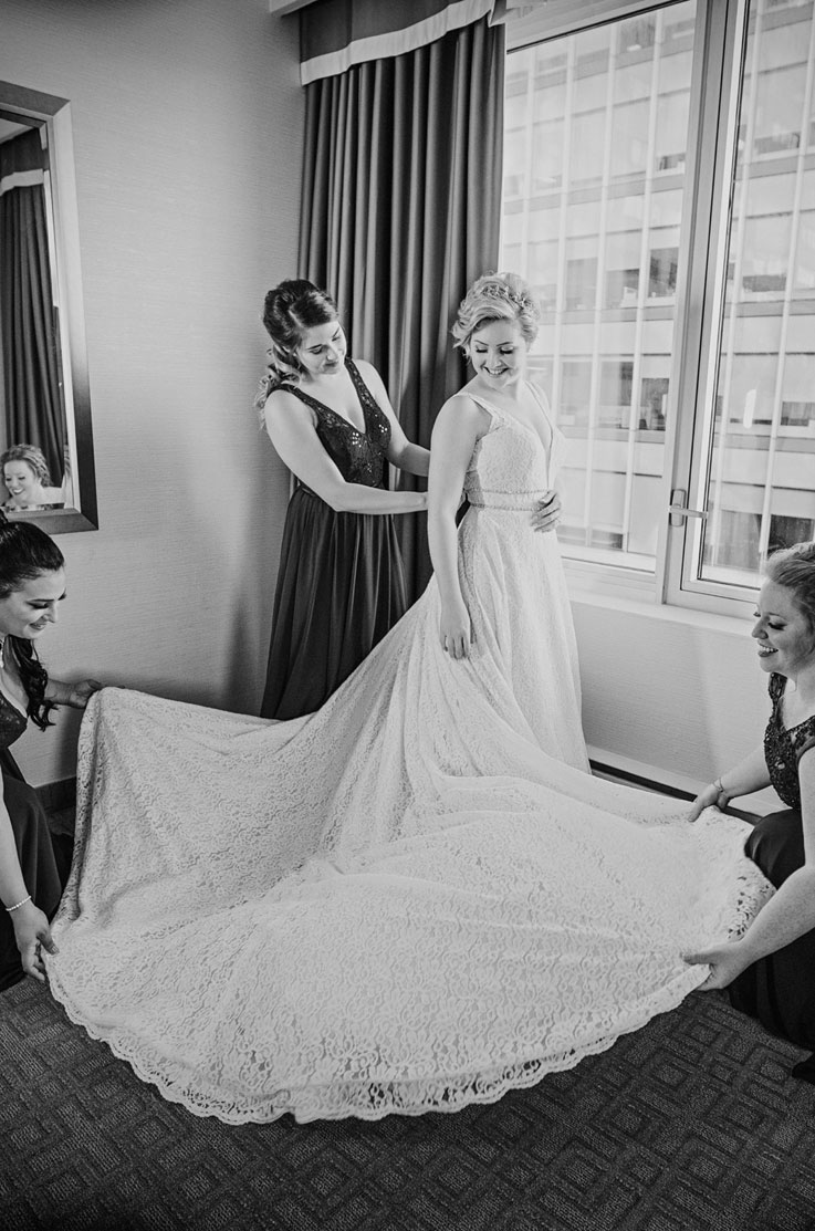 Bride in a lace wedding dress with a plunging neckline, stands by a window in a bridal suite with bridesmaids helping.