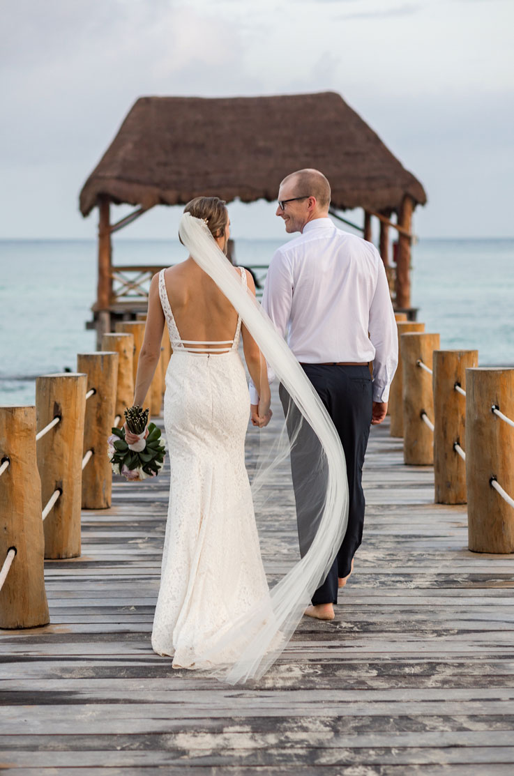 Bride in a fit and lace wedding dress with a low back and flowing veil, walks hand in hand with groom on a wooden pier.