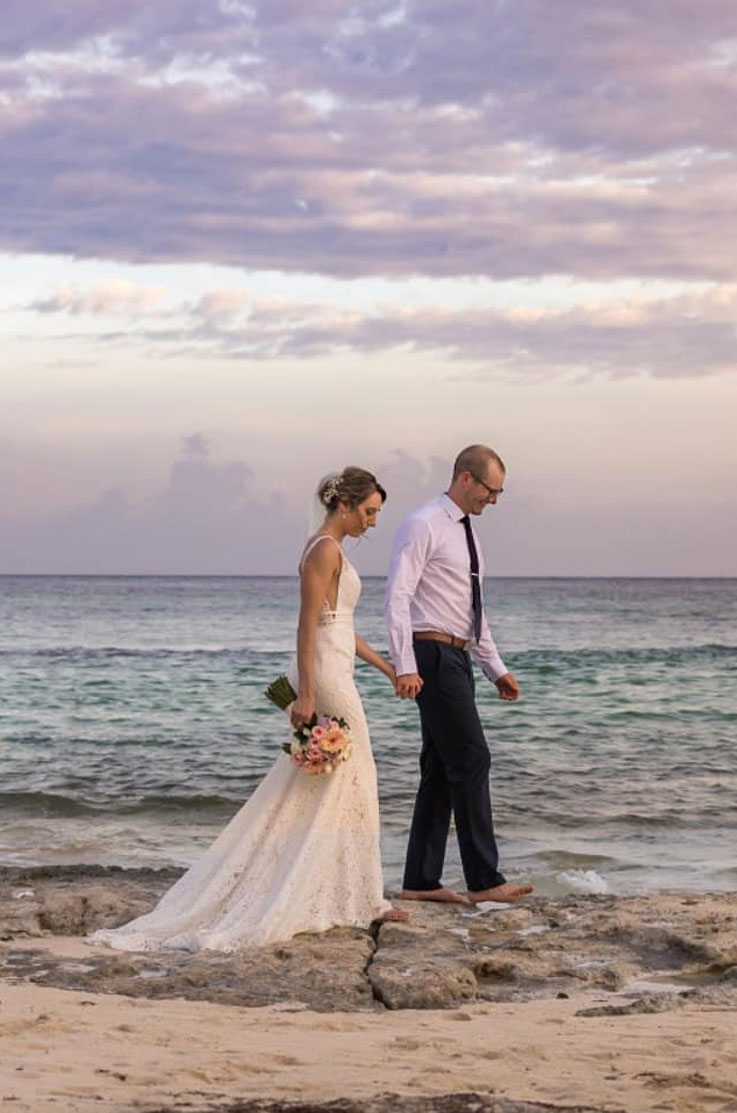 Bride in a sleeveless lace gown with double bands at the waist, holds hands and walks with her groom on a rocky shoreline.
