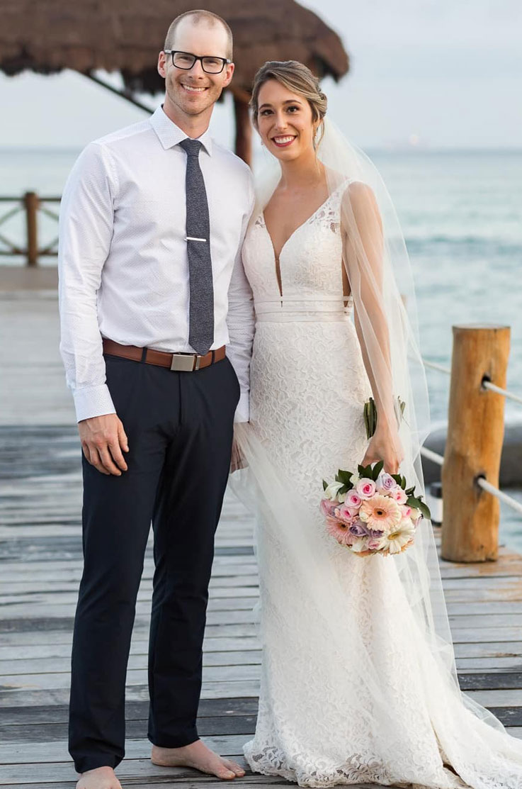 Bride in a V-neck lace gown with a long veil, smiles standing next to groom, and is holding a bouquet on a pier over the ocean.
