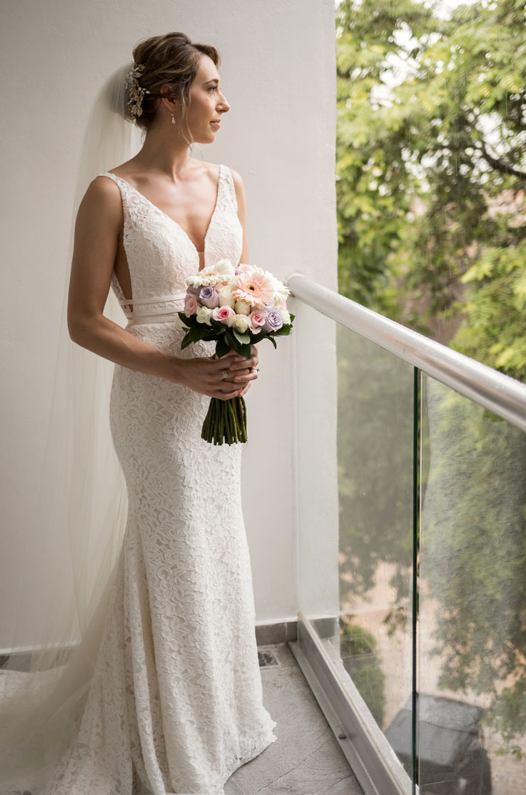 Bride in a fit and flare lace gown with a V-neckline, veil and thin bands on the bodice, is holding a bouquet on a balcony.