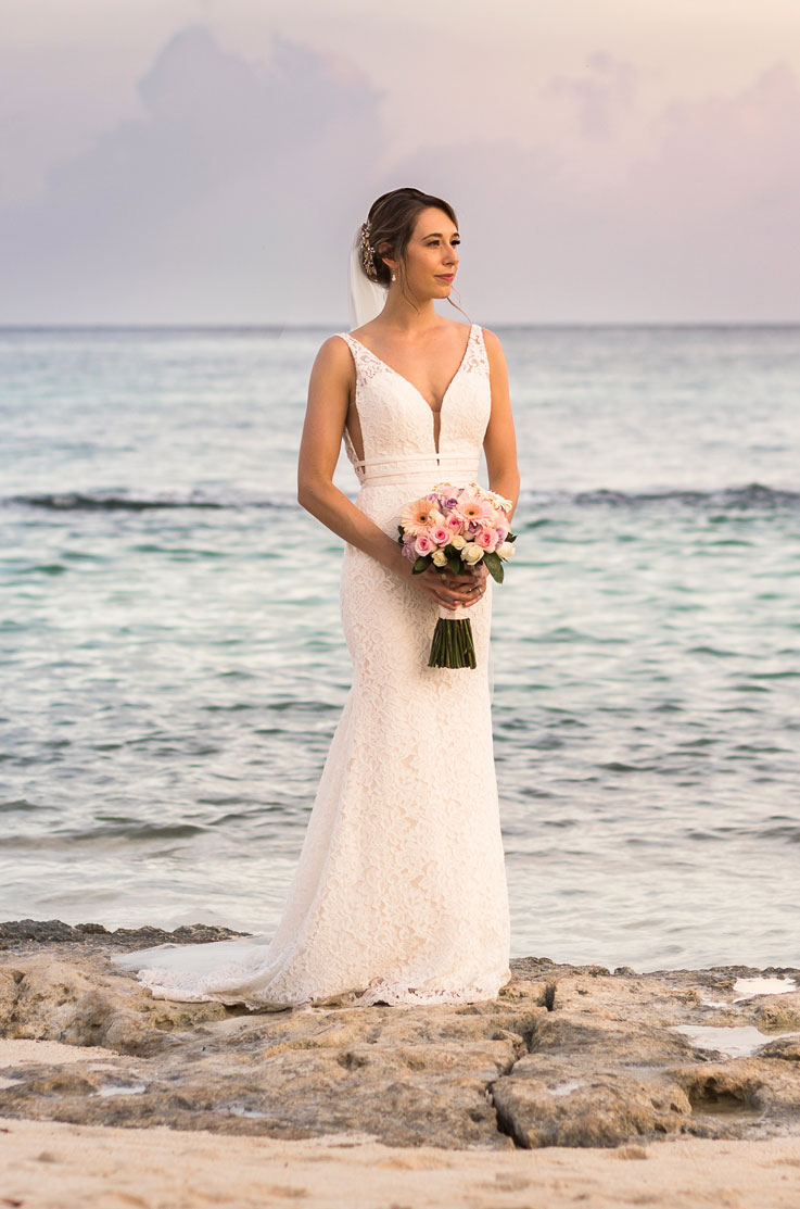 Bride in a lace fitted gown with a V-neck and thin double bands, holding a bouquet on a rocky shore as the sun sets.