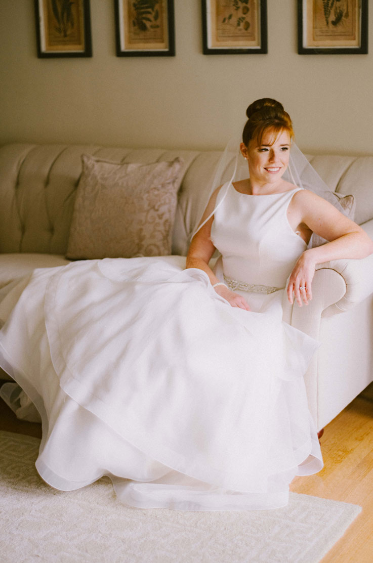 Bride in a sleeveless satin bodice wedding dress with a full skirt, beaded belt and veil, leaning on a light colored sofa.