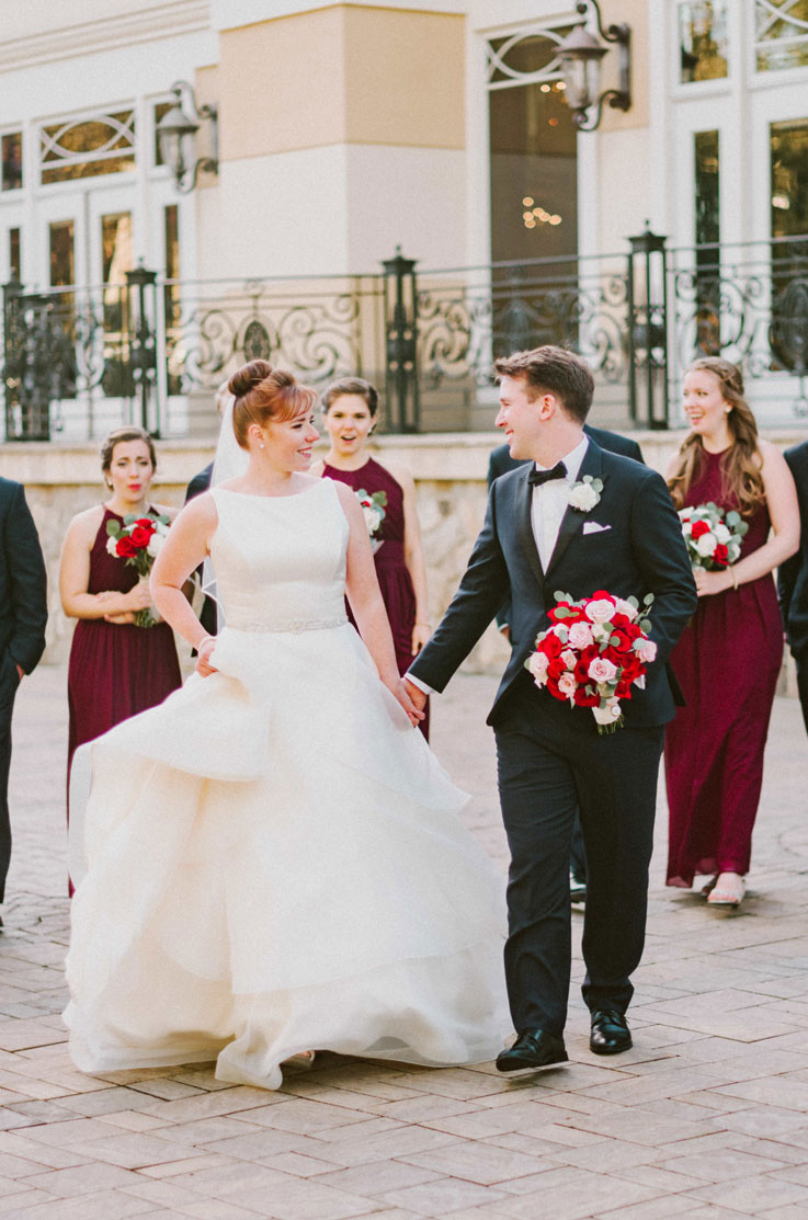 Bride in a satin gown with a layered skirt and belt, holds hands and smiles at her groom holding a pink and red bouquet.