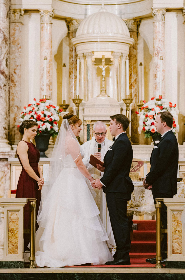 Bride in an organza ballgown with a layered skirt, a beaded belt and veil, is facing groom holding hands at a church altar.