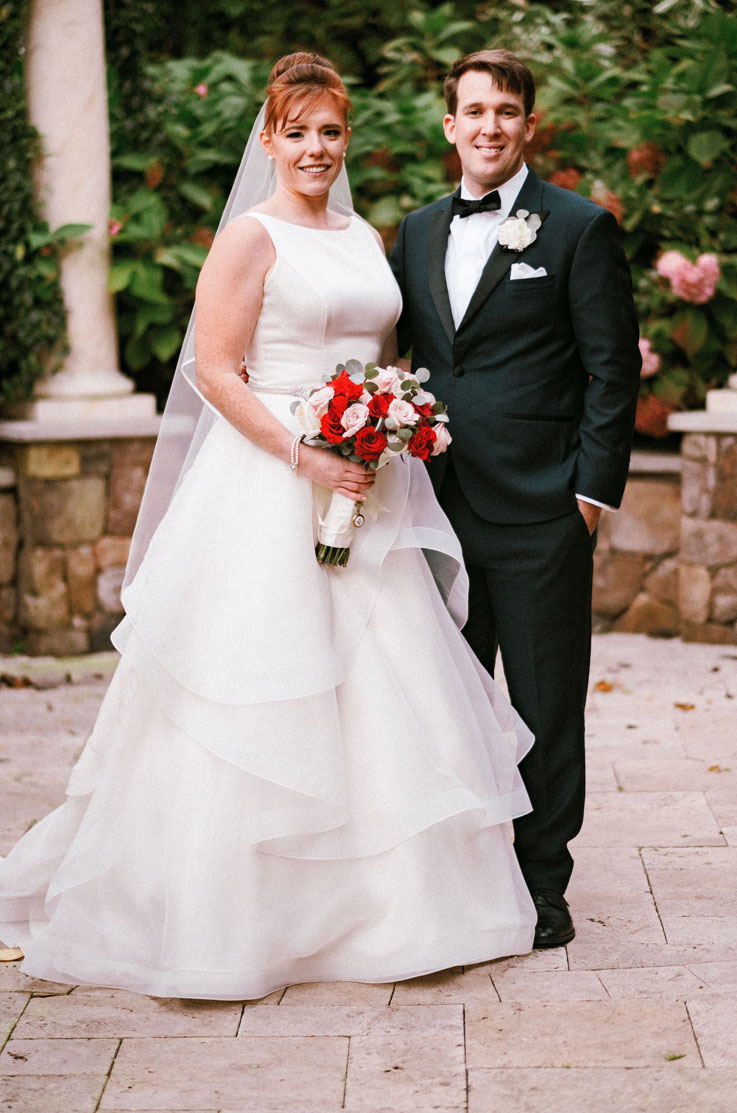 Bride poses in a satin bodice gown with a beaded belt, veil and a layered organza skirt, holding a bouquet next to her groom.