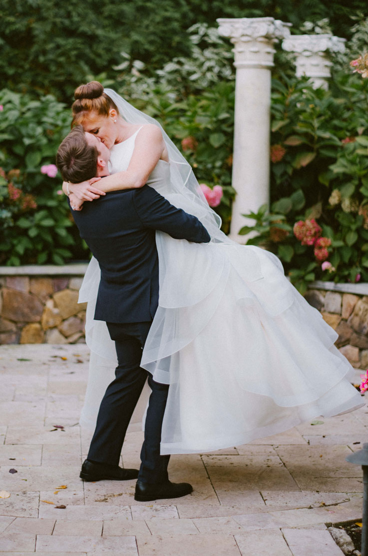 Groom lifts his bride in a sleeveless organza gown with a satin bodice and veil, and they kiss on a stone terrace.