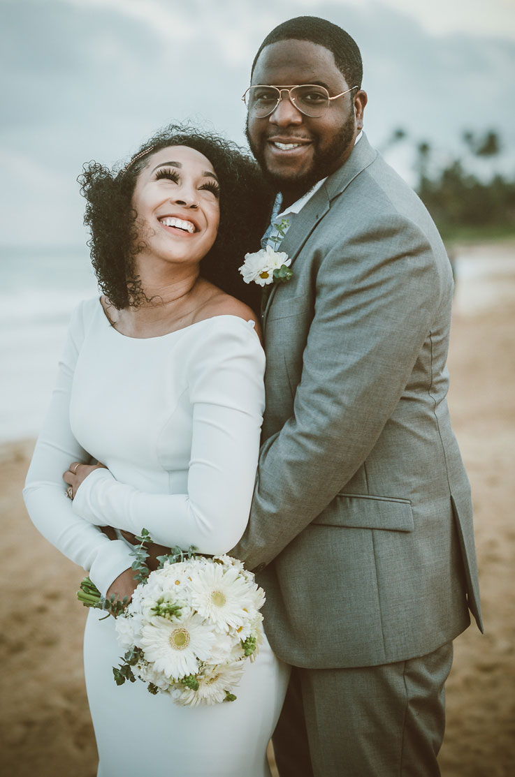Bride in a crepe long sleeve wedding dress with a bateau neckline, holding a bouquet and smiles at groom on a sandy beach. 