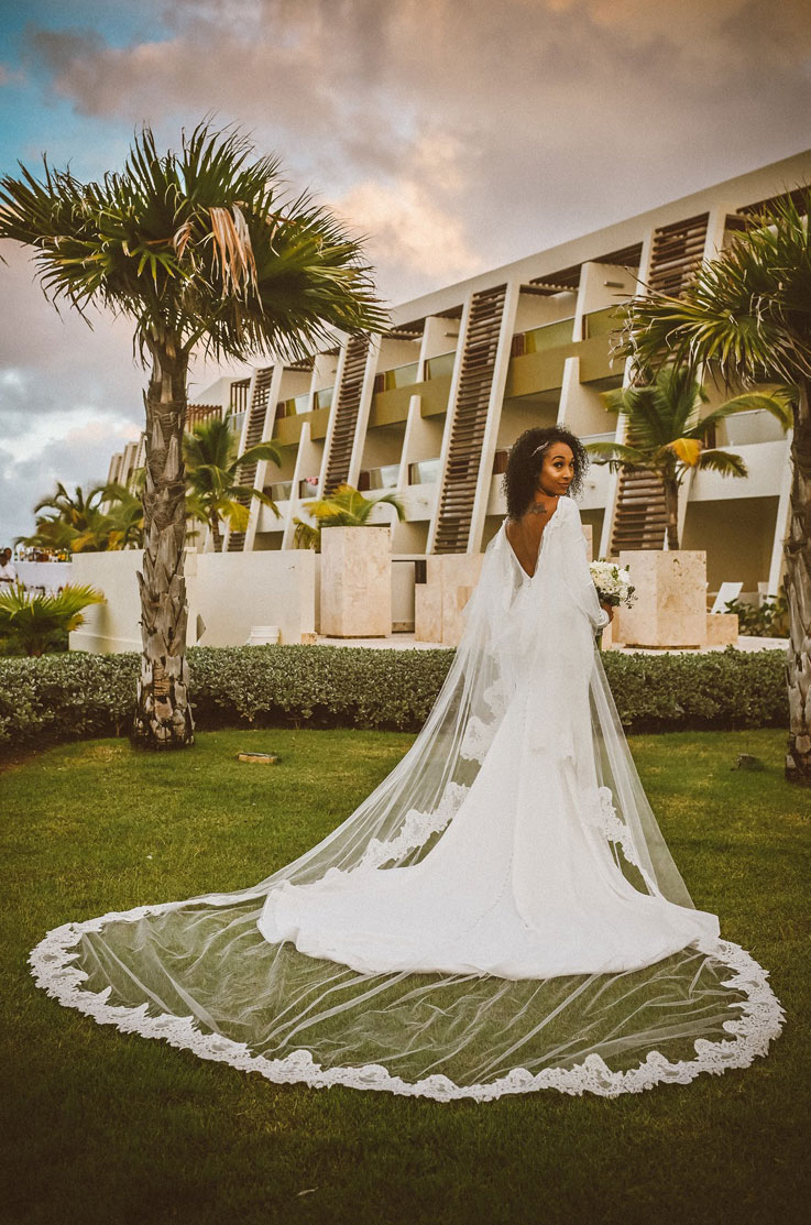 Bride in a crepe fit and flare gown with a cape, sleeves and a low back, looks over her shoulder in a tropical resort. 