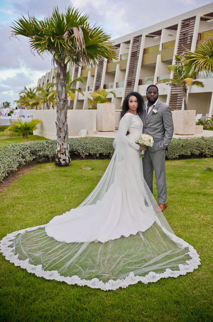 Bride poses next to groom, in a bateau neck crepe wedding gown with sleeves, holding a bouquet and standing by palm trees.