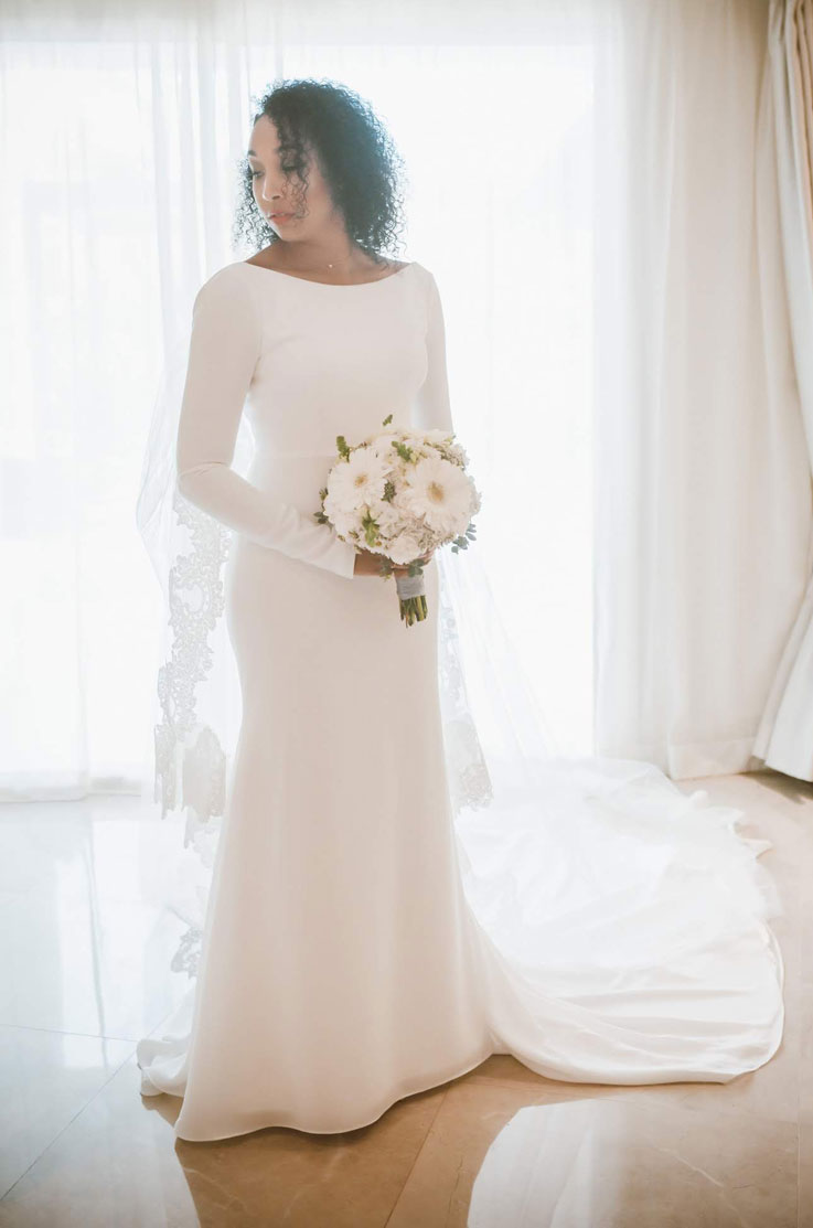Bride poses by a bright window with a bouquet, in a crepe fit and flare gown with sleeves, a bateau neckline and a lace cape.