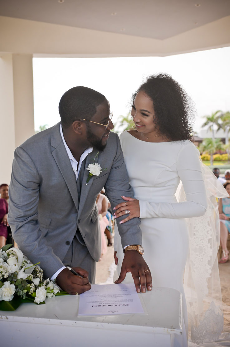 Bride smiles in a crepe long sleeve gown with a cape and a bateau neckline, smiling with groom and signing marriage papers.