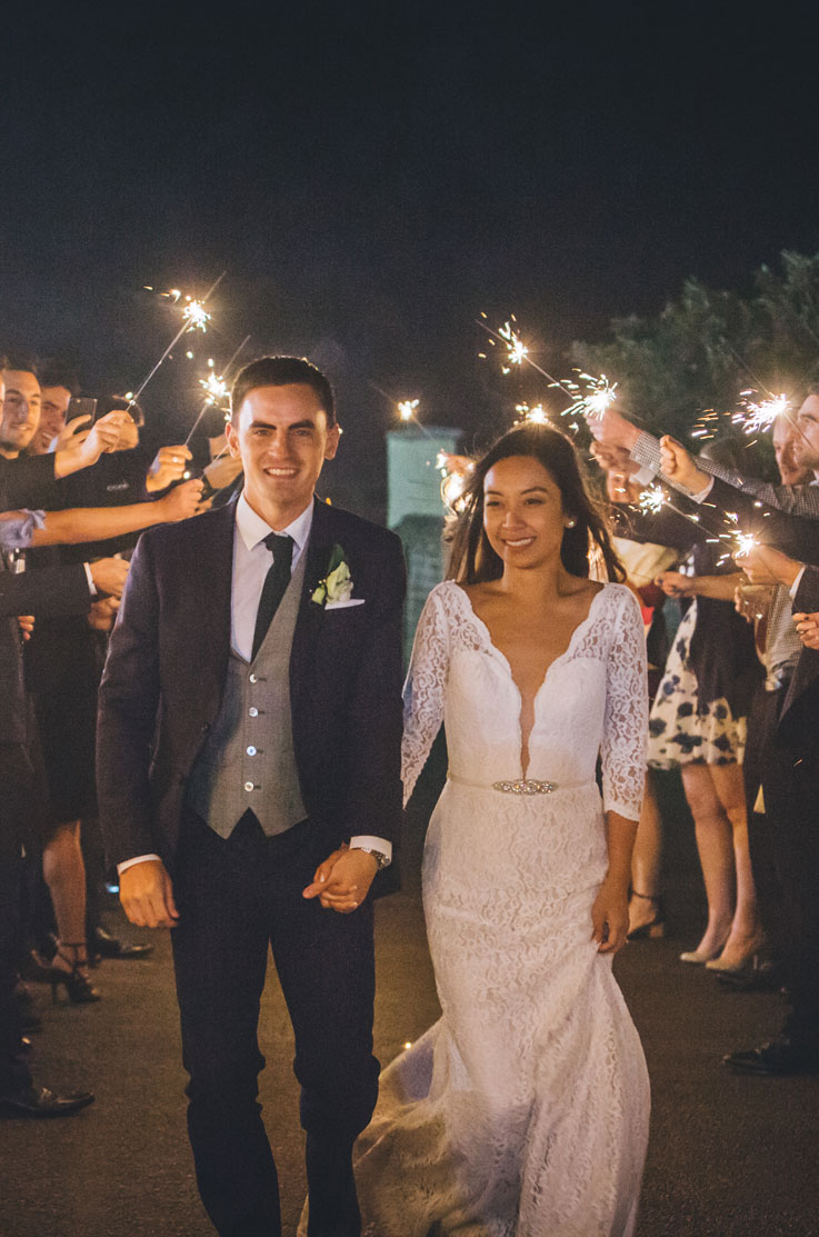 Bride in a lace wedding gown with a plunging neck and a belt, walks hand in hand with her groom through a sparkler exit. 