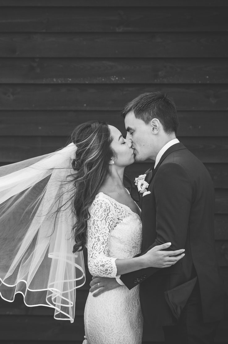 Close-up of the bride in a lace long-sleeve wedding dress with a flowing veil, kissing groom and taken in black and white.