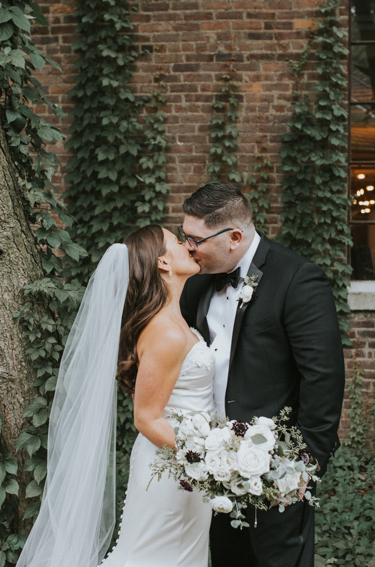 Bride in a strapless fit and flare gown with a veil, holds a bouquet and kisses groom in front of an ivy-covered brick wall.